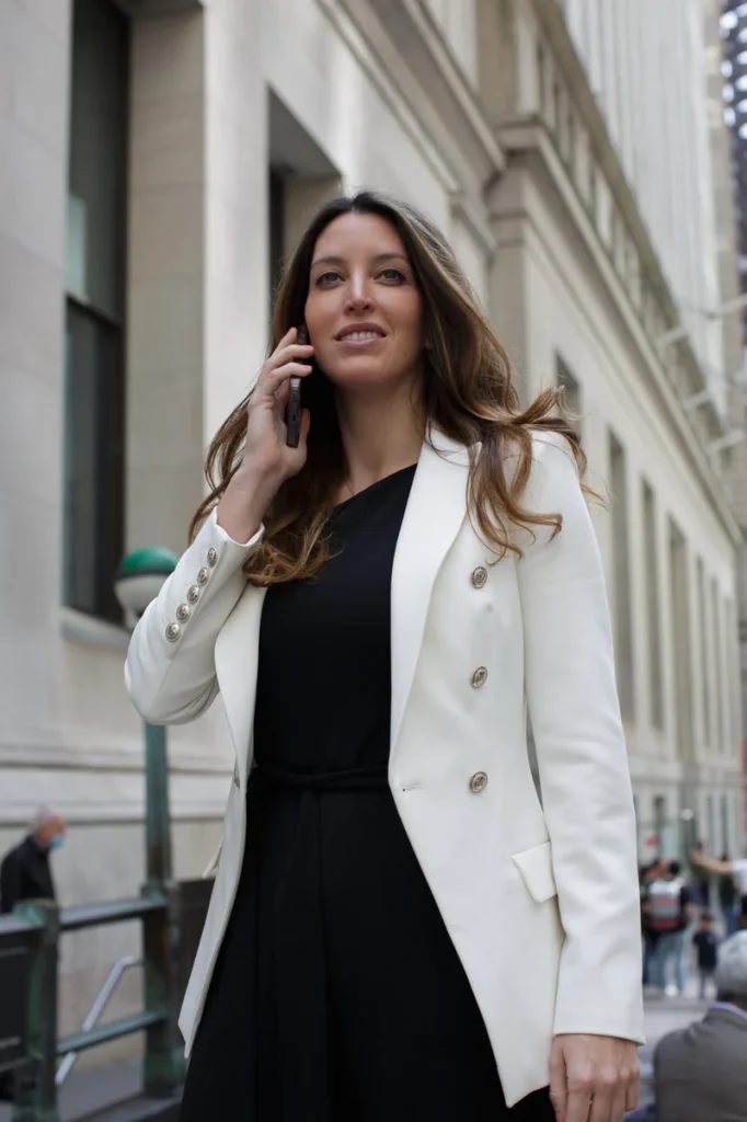 Mujer con blazer blanco y vestido negro hablando por teléfono en una calle de edificios de piedra
